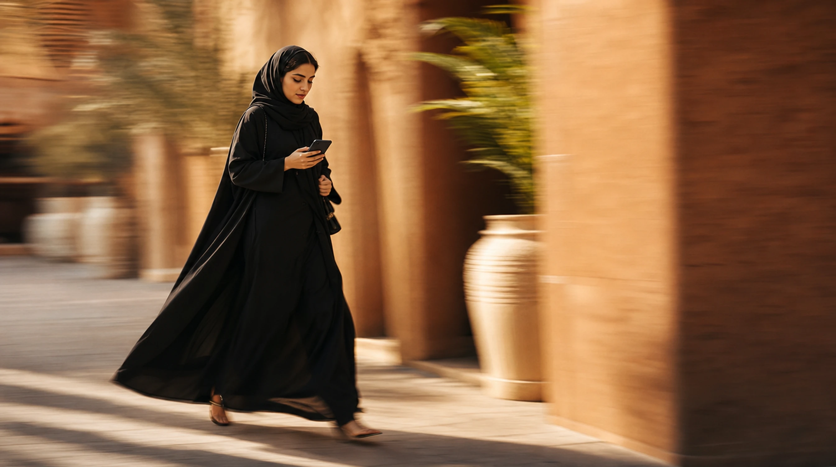 A Saudi woman in a modern abaya walking and browsing her phone in warm light